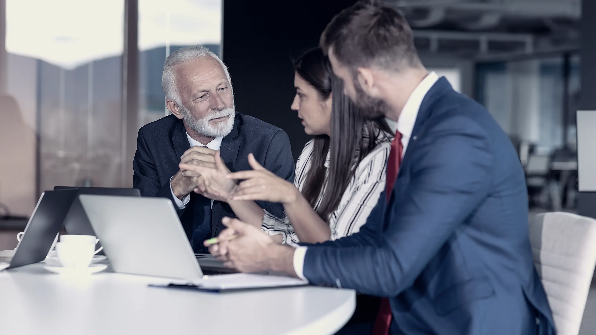 A senior adviser in discussion with two executives across a meeting table.