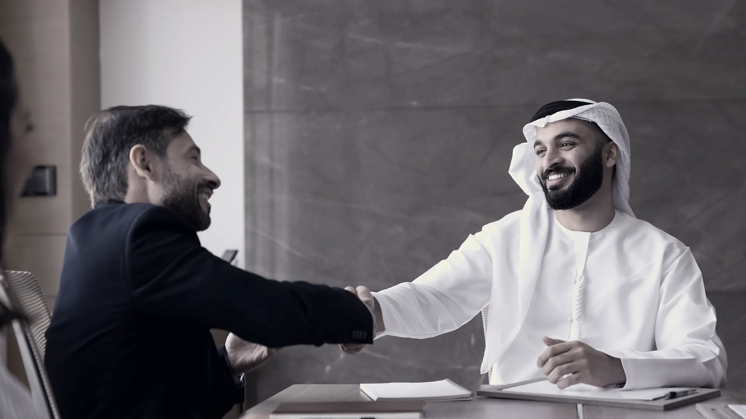 A Gulf executive and a Western executive shaking hands across a boardroom table.